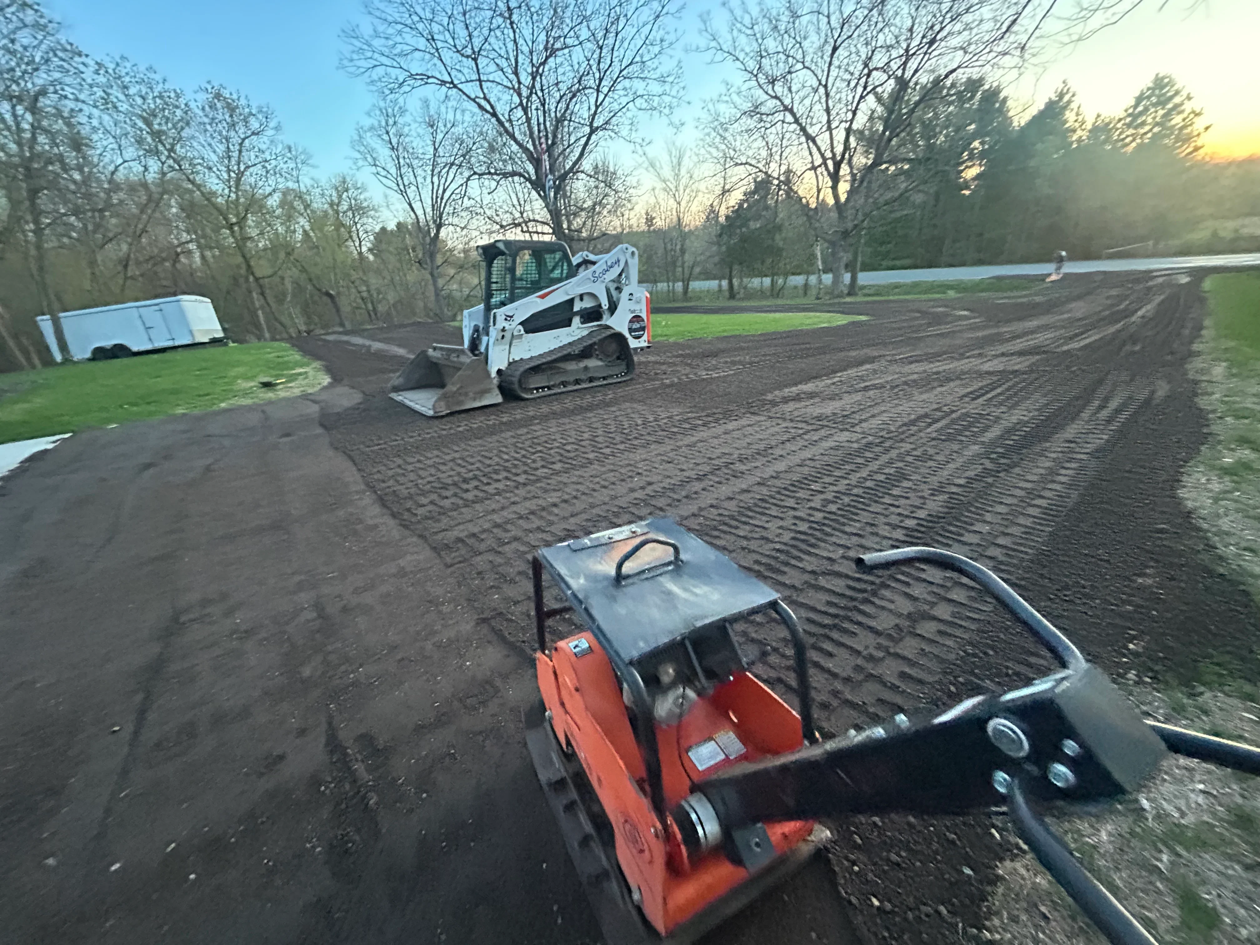 Compact track loader grading driveway with plate compactor in foreground on prepared site