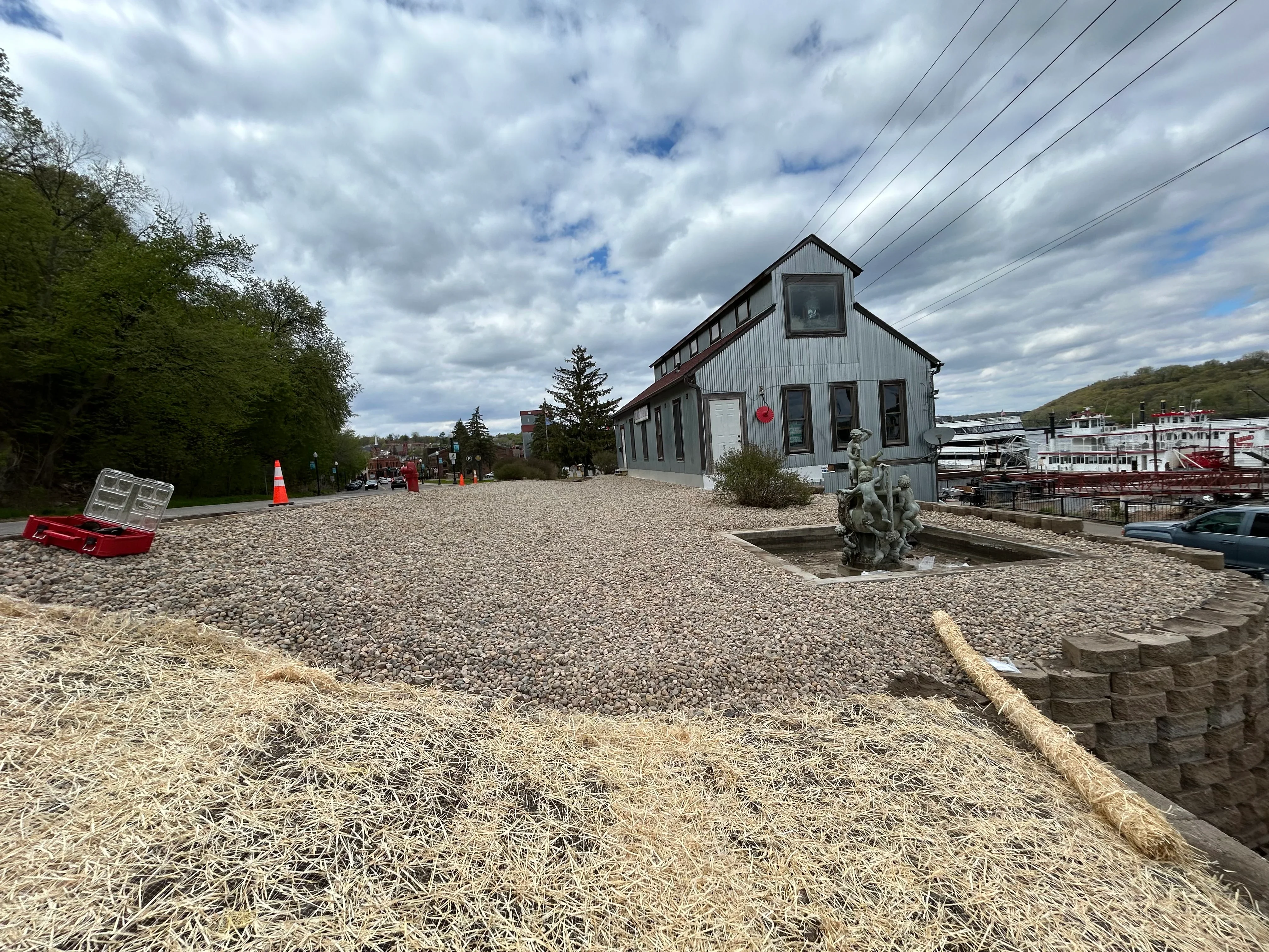Gravel-filled excavation site with prepared foundation near riverfront building and docked boats