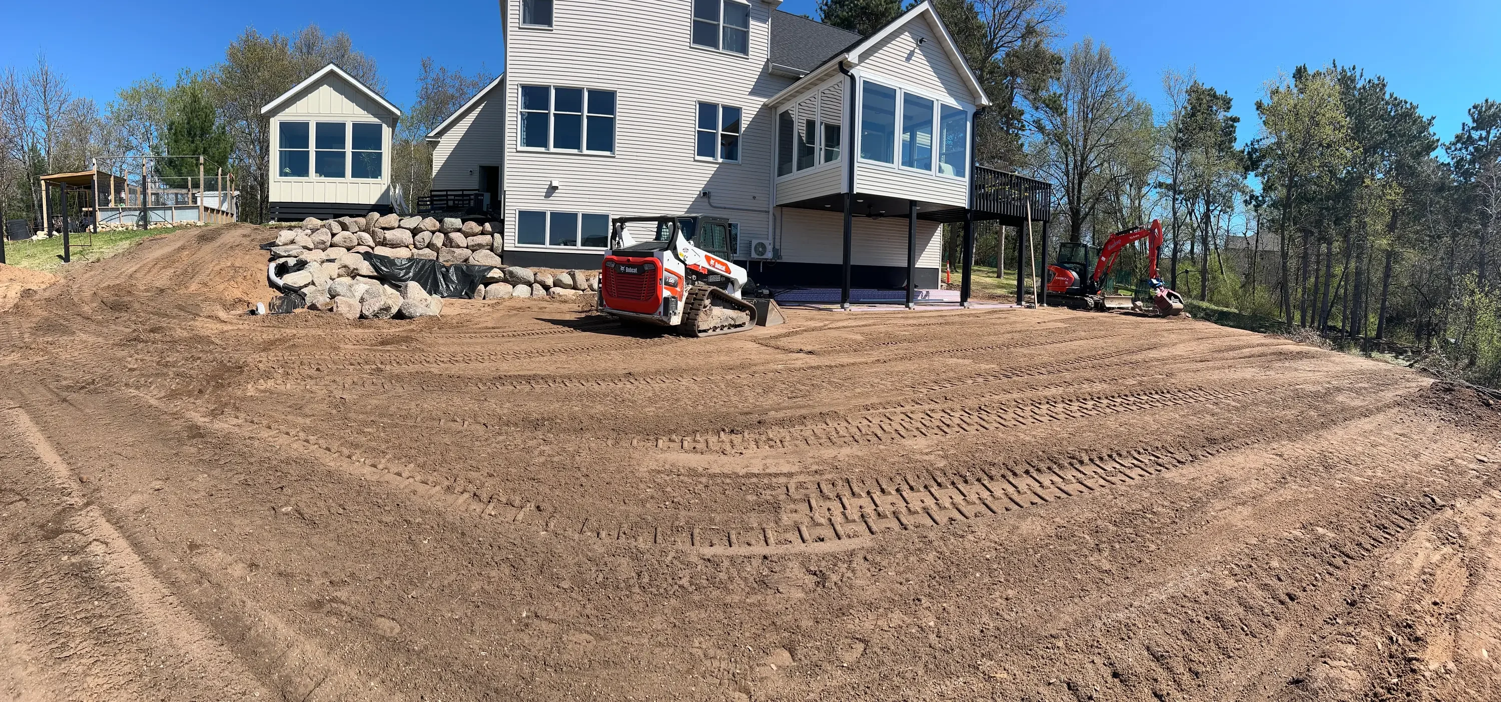 Excavated residential driveway with graded soil, boulders, and modern elevated home under construction