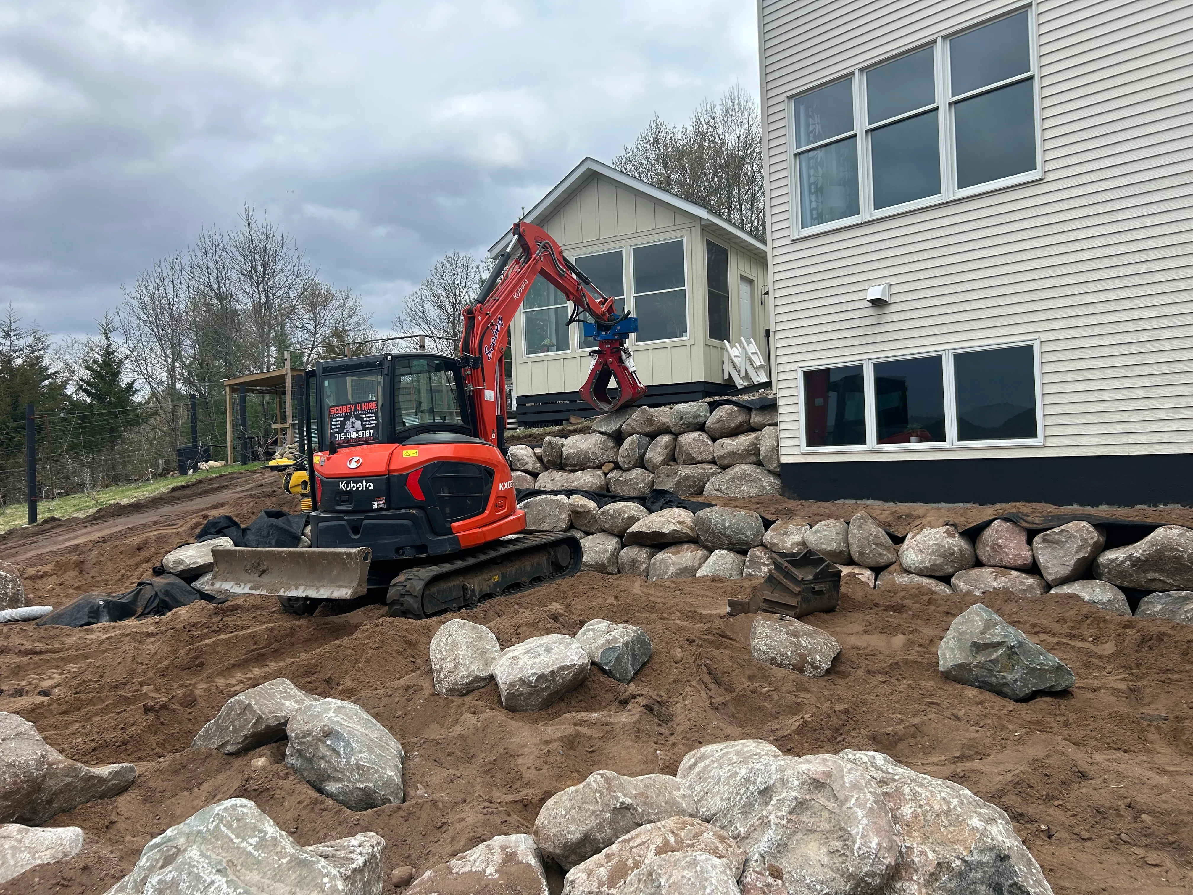 Red Kubota excavator placing large boulders next to residential home with vinyl siding