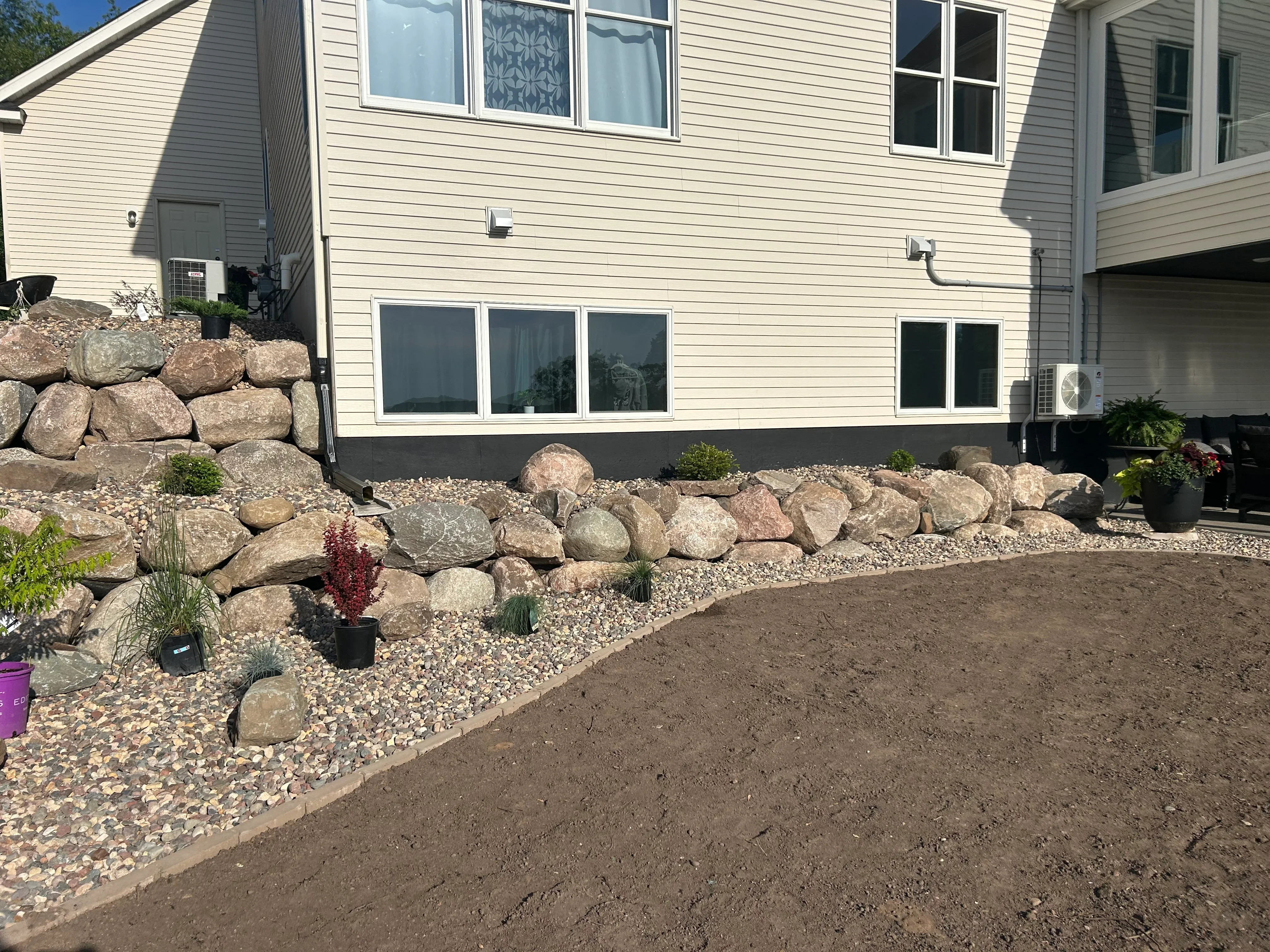 Cream vinyl-sided home with new boulder retaining wall, gravel landscaping, and prepared grading