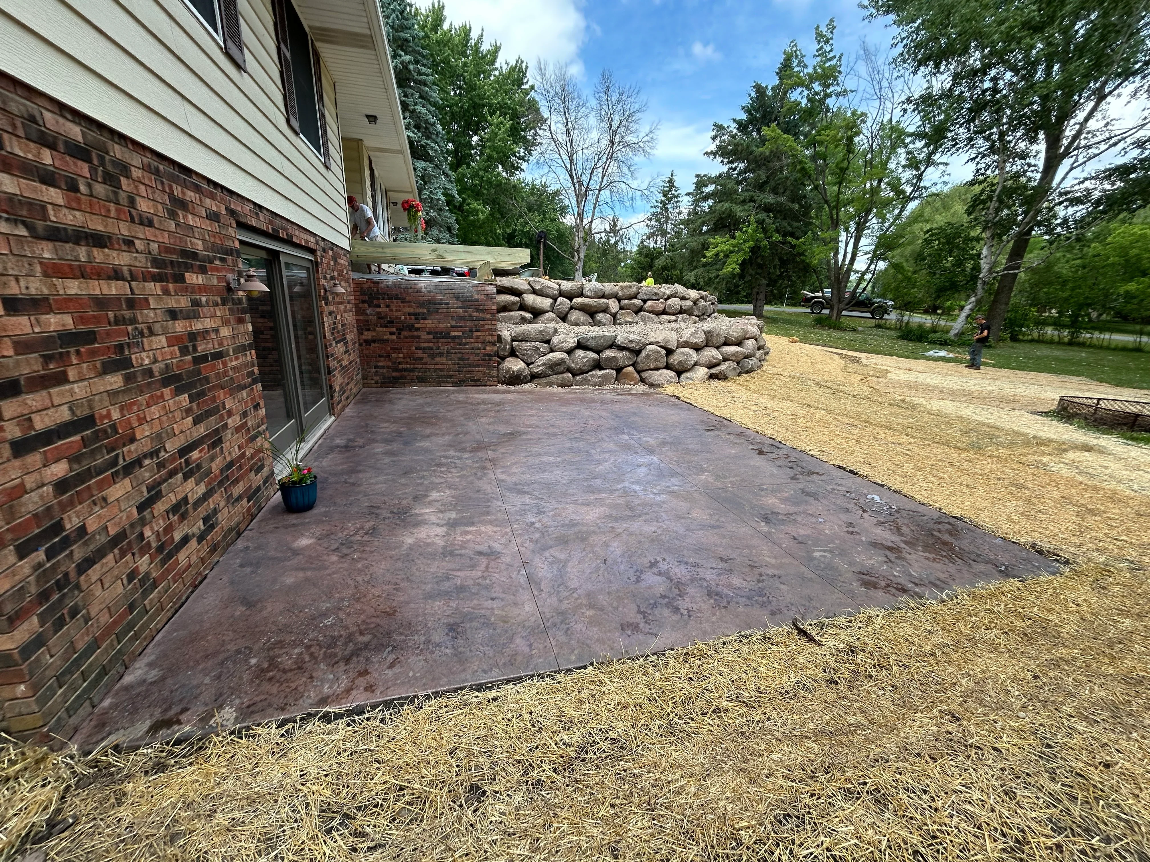 Excavated patio pad with straw erosion control and boulder retaining wall beside brick home